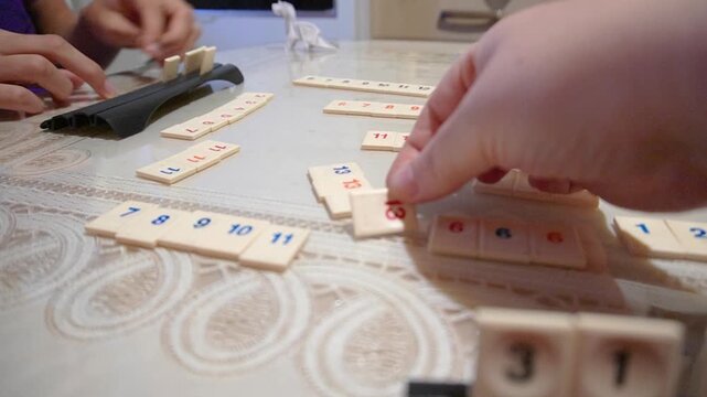 Family playing Rummikub board game at table. Hands arranging numbered tiles with racks, indoor home environment, tabletop strategy activity.