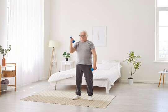 Focused gray haired senior man standing in bedroom and lifting dumbbells. Active pensioner doing fitness exercise in morning at home, working out with weights to improve mobility, muscle and health.