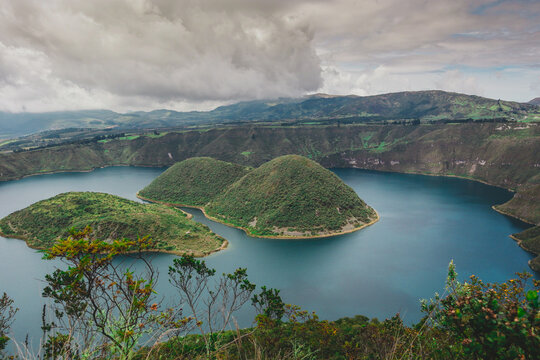 A stunning panoramic view of Laguna de Cuicocha, a deep blue crater lake within the Cotacachi Volcano. The image highlights its two distinct volcanic islands and the steep, lush caldera walls under a