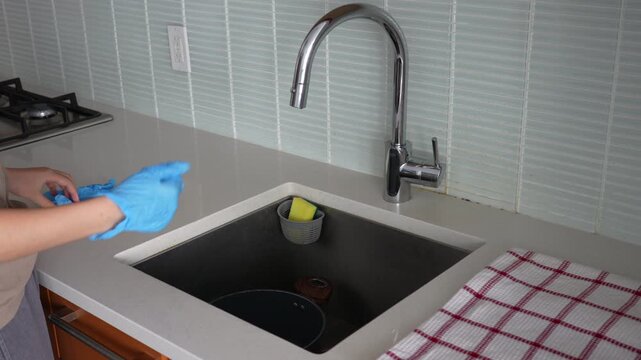 A person put on rubber gloves, turn on water, and wash yellow sponge. The clean, modern kitchen creates a fresh, hygienic mood. Use for household, hygiene, or daily routine themes