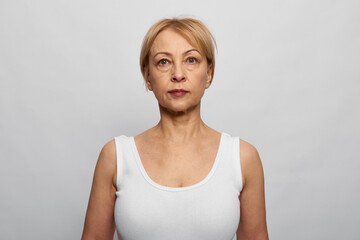 Mature woman with short blonde hair wearing white tank top stands against neutral gray background. Serious facial expression, natural look, portrait concept for age and confidence. © SHOTPRIME STUDIO