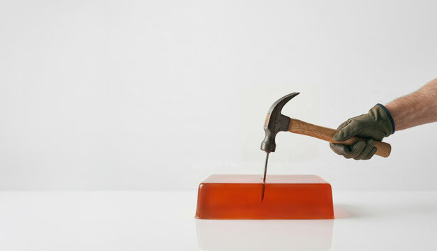 A gloved hand uses a hammer to drive a nail into splashing orange gelatin, illustrating overcompensation and excessive force against a white background.