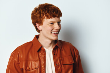 Obraz premium Young man with red hair wearing brown leather jacket smiling and looking aside on white background. Casual style portrait with happy expression and natural lighting.