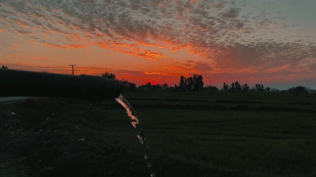Water Splashing Over Green Field At Vibrant Sunset With Trees Road and Dramatic Golden Hour Sky
