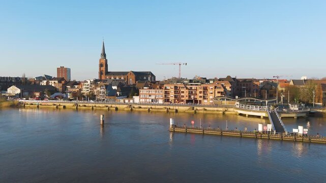rippling blue water fronts boom waterfront floating jetty pedestrian ramp backed by brick houses tall gothic church spire beneath clear sky riverside town urban landscape residential area 