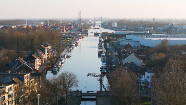 white yachts line klein willebroek calm canal brick townhouses pale warehouses extending shipping containers metal lifting bridges soft morning haze toward near riverside town urban landscape 