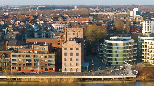historic pale brick industrial building arched windows sits among wooden clad apartments white and green residential blocks facing riverside promenade lined reeds metal 