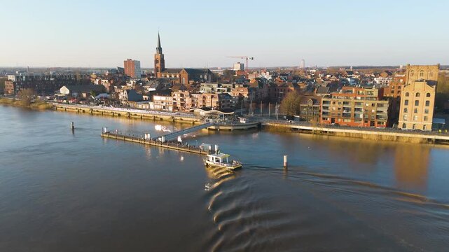 small ferry glides across river floating dock leaving rippling wake under warm light brick buildings tall church spire along waterfront promenade boom toward broad 