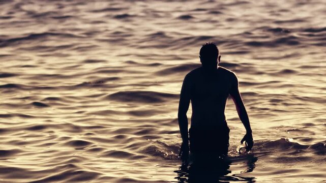 Silhouette diver person walking in shallow water during scenic sunset holding goggle mask