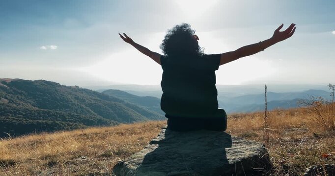 Hiker woman sits on rock with outstretched arms illuminated by the golden setting sun against mountainous landscape, concept of success and victory