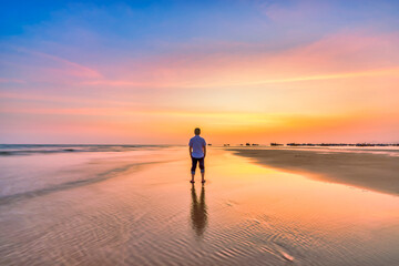 The silhouette of a man stands alone on a wide sandy beach while watching a vibrant sunset over the ocean. © huythoai