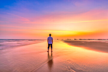 The silhouette of a man stands alone on a wide sandy beach while watching a vibrant sunset over the ocean. © huythoai