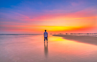 Fototapeta premium The silhouette of a man stands alone on a wide sandy beach while watching a vibrant sunset over the ocean.