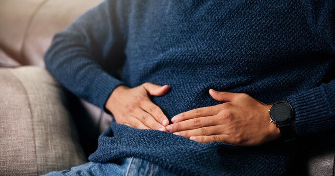 Man, hands or stomach ache with pain for indigestion, constipation or diarrhea on sofa in home. Closeup, male person or sore belly with gastrointestinal reflux for virus or bladder infection in house