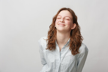 Fototapeta premium Young woman with curly hair smiles with closed eyes wearing white shirt on light background. Concept of happiness, relaxation, and positive emotions in casual lifestyle.