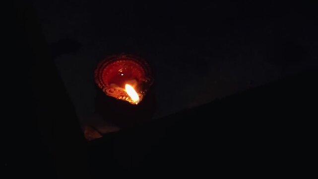 A flickering diya, an oil lamp, glows warmly in the darkness. The orange flame dances within its clay base, casting a soft, golden light. 