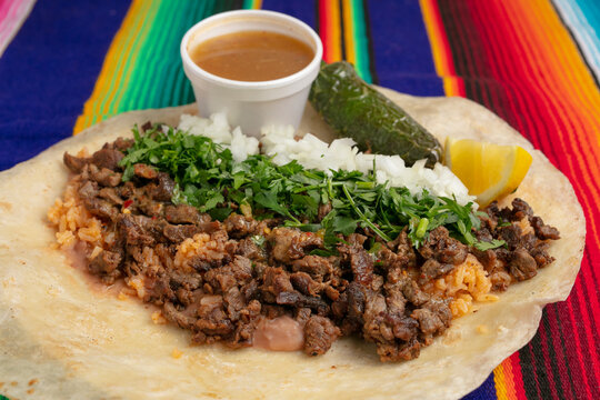 Close up shot of an open carne asada burrito with cilantro, onion, lime, salsa, rice and jalape&ntilde;o on a flour tortilla and traditional Mexican tablecloth 