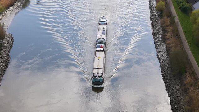 straight elongated inland cargo vessel along river centerline teal bow cleaving silvery surface mirrored wave patterns spreading stone armored banks walkway toward down drone shot 