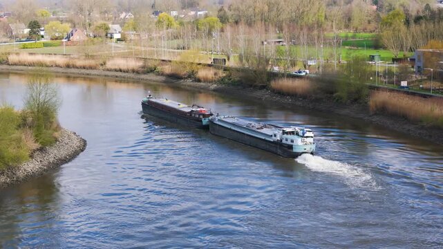 coupled inland motor vessel rounds gentle river bend white wheelhouse leading barge foamy stern wash trailing bordered by reed lined banks roadside traffic scattered suburban homes 
