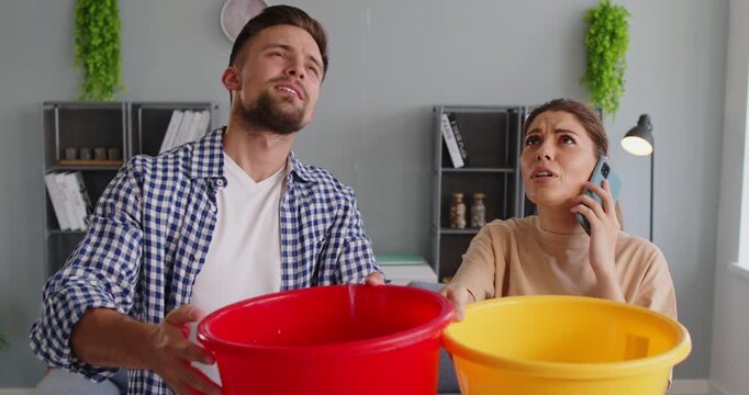 Couple holding buckets under ceiling leak. At home, they catch water falling from roof while calling for repair to reduce damage and stains. Leakage, home maintenance and emergency response.