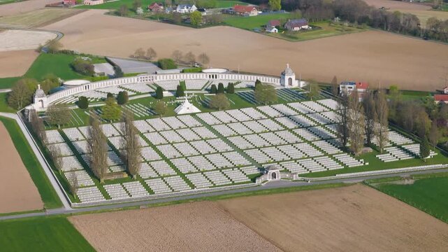 vast military tyne cot cemetery within open farmland has perfectly aligned white headstone rows manicured lawns divided by paths converging central cross sacrifice tall trees cast shadows; 