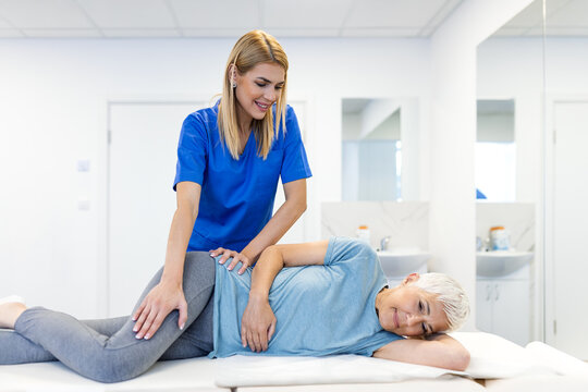 Professional female therapist performing manual hip therapy on a senior woman in a bright clinic. Specialist assisting an elderly patient with lower back and pelvic stability exercises.