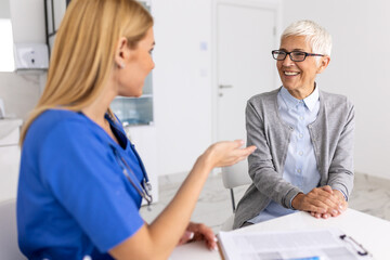 Fototapeta premium Happy senior woman smiling during consultation with female doctor in blue scrubs friendly medical professional providing geriatric care and emotional support in bright hospital office.