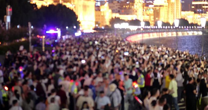 Blurred view of huge crowd of people walking along Huangpu River embankment on holiday night; moving mass of tourists resembles river, with bright flashing police beacons controlling human traffic