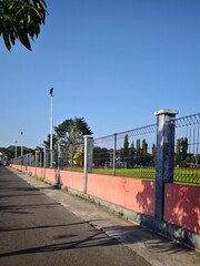 A long perspective of a metal wire fence with pink concrete base surrounding a green soccer field under a clear blue sky