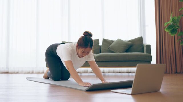Young Asian woman stretching yoga workout on exercise mat while online training class with computer laptop in living room