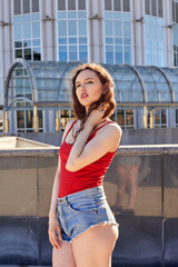 Obraz premium A young woman with long dark hair is standing in front of a city building. She is wearing a red top and denim shorts. In the background is a modern cityscape in daylight.