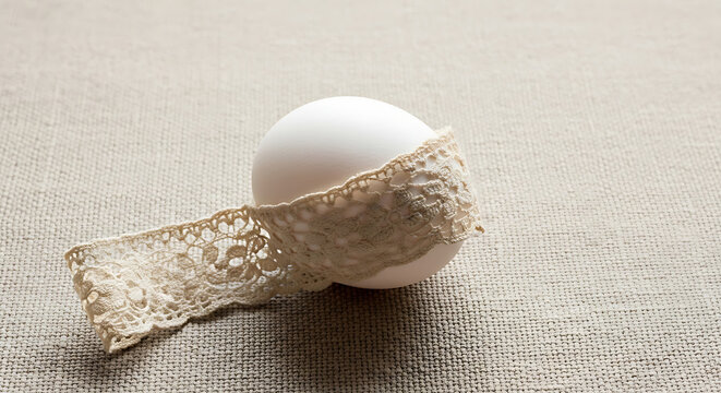 A single white egg with delicate lace ribbon wrapped around it, resting on a textured fabric surface.