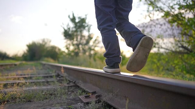 Close up of boy feet walking on railroad track. Child in jeans balancing on metal rail at sunset. Legs move forward on railway line. Kid in sneakers balances on railroad track step by step.