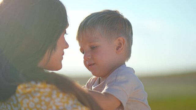 Happy mother kisses little son on forehead outdoors. Family enjoys sunny summer day with love. Child laughs and shows tongue playfully. Happy mom and kid hug with love in nature park close up.