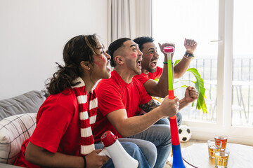 Fototapeta premium Diverse group of young adult friends wearing red jerseys, cheering and celebrating a goal during a live soccer match at home
