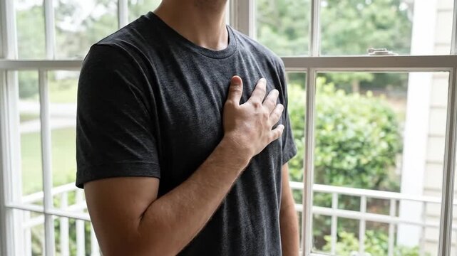 Man placing hand over heart, conveying sincerity, respect, or a pledge indoors by a window with natural light.