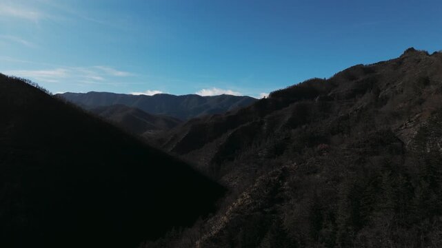 Cinematic aerial dolly in across rugged mountain slopes and a deep shadowed valley with sparse forest under a clear blue sky.