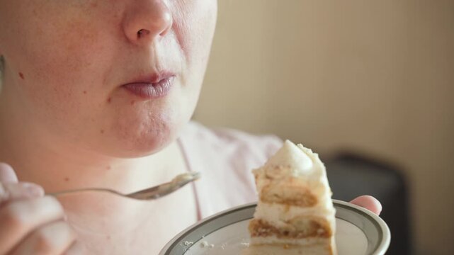 Full woman eats and chews cake slice at home, closeup of face with mouth. Overweight woman eats sweet dessert with spoon, eating behavior problem.