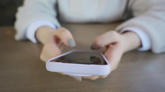 Female hands actively type and scroll on a purple smartphone over a blurred wooden desk. Concept of modern digital communication, online social networking, and wireless lifestyle.