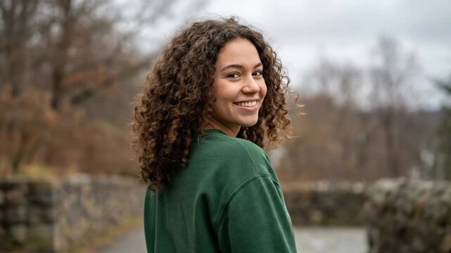 Smiling young woman with beautiful curly hair looks over her shoulder outdoors, radiating positive energy and natural charm.