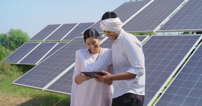 Indian mature adult woman hold tab look screen visit farm field do work check record data at rural area ground place female talk speak share eco friendly solar panel power plan idea to young male