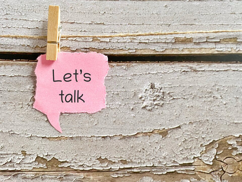 Pink speech bubble sticky note with the phrase lets talk clipped on a string with rustic wooden background representing communication conversation and connection concept.