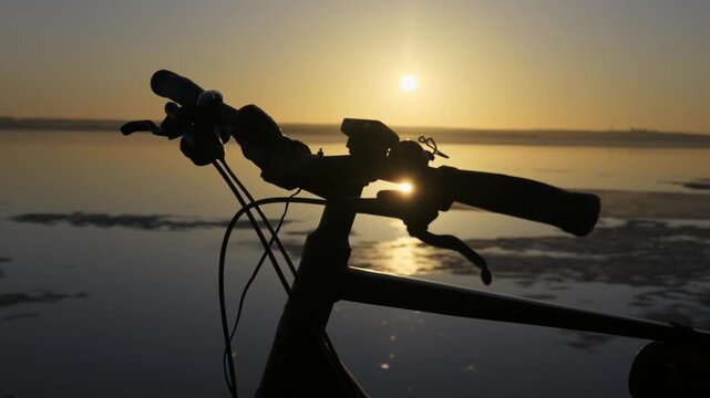 A silhouette of bicycle handlebars stands against a glowing golden hour sunrise reflecting brightly across a calm lake. Concept of morning exercise, outdoor adventure, and nature.