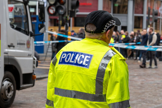 Police officer in a high-visibility jacket at a cordoned city street scene with an emergency vehicle and a crowd.