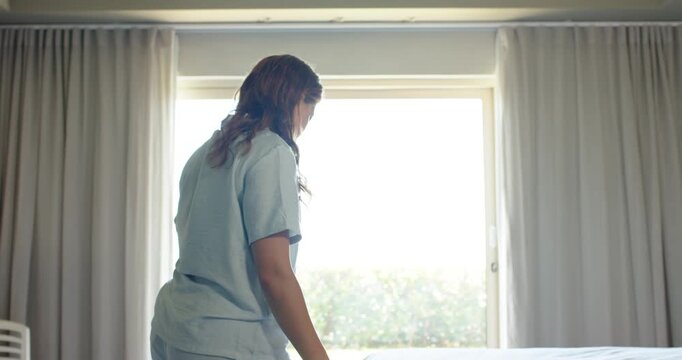 African American woman in pjs by bright window pulling tucking smoothing duvet, making bedroom bed
