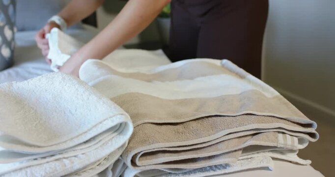 Folding woman in dark brown outfit stacking towels on bed, laundry basket, wristwatch, copy space