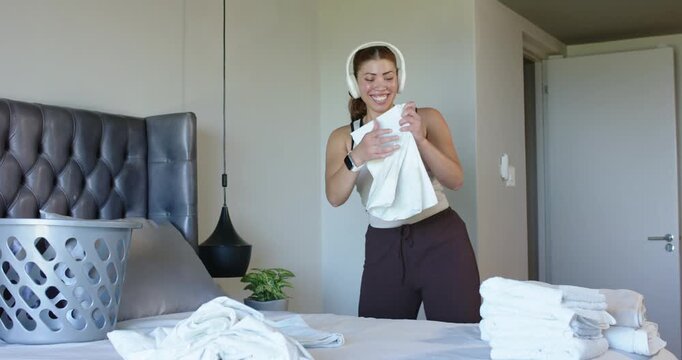 African American woman lifting towel, folding on bed with basket, headphones watch creating stack