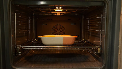 Fototapeta premium Cozy Kitchen Scene With Steaming Casserole. Golden Edged Casserole In Oven On Metal Rack Emitting Heat. Intimate Closeup Of Baking Ceramic Dish With Steam And Glowing Edges In Kitchen