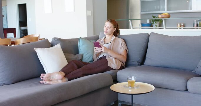 African American woman checking pink phone, leaning on gray couch with side table glass, copy space