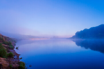 Fototapeta premium Morning mist covers a river in a quiet area as the sun starts to rise over the trees and water surface creating a soft glow
